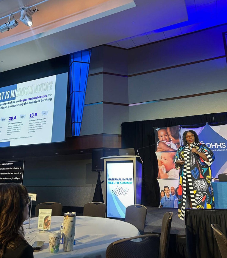 Presenter speaking on stage at a conference with slides displayed nearby.